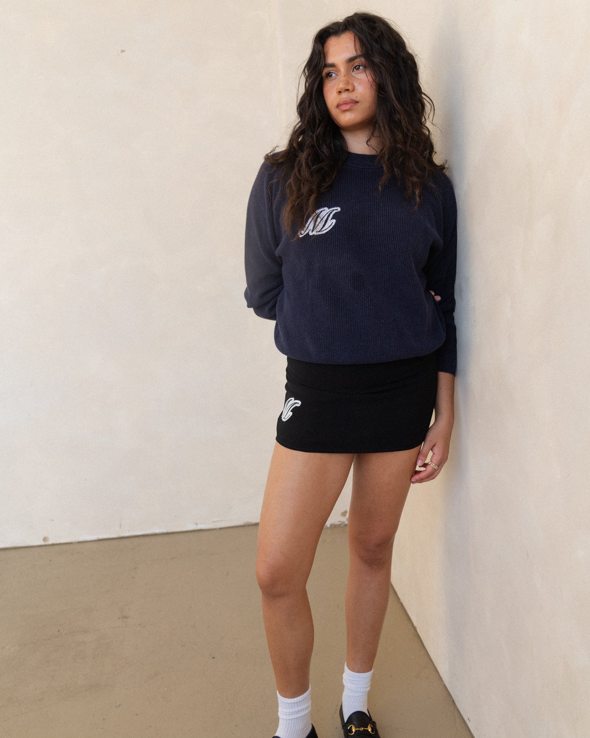 Woman wearing a navy sweatshirt and a black skirt, both monogrammed with an M on the model's right side, against a beige wall.