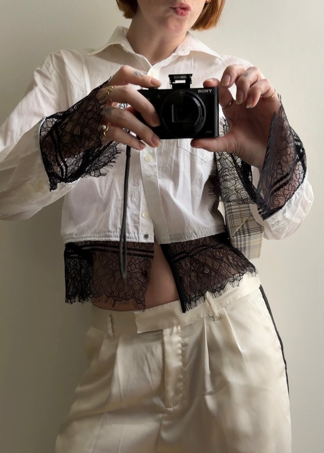 Person wearing a white shirt with black lace sleeves and holding a camera.