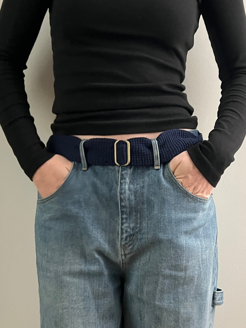 Person wearing a black long-sleeve top and blue jeans with a belt, standing against a plain background.