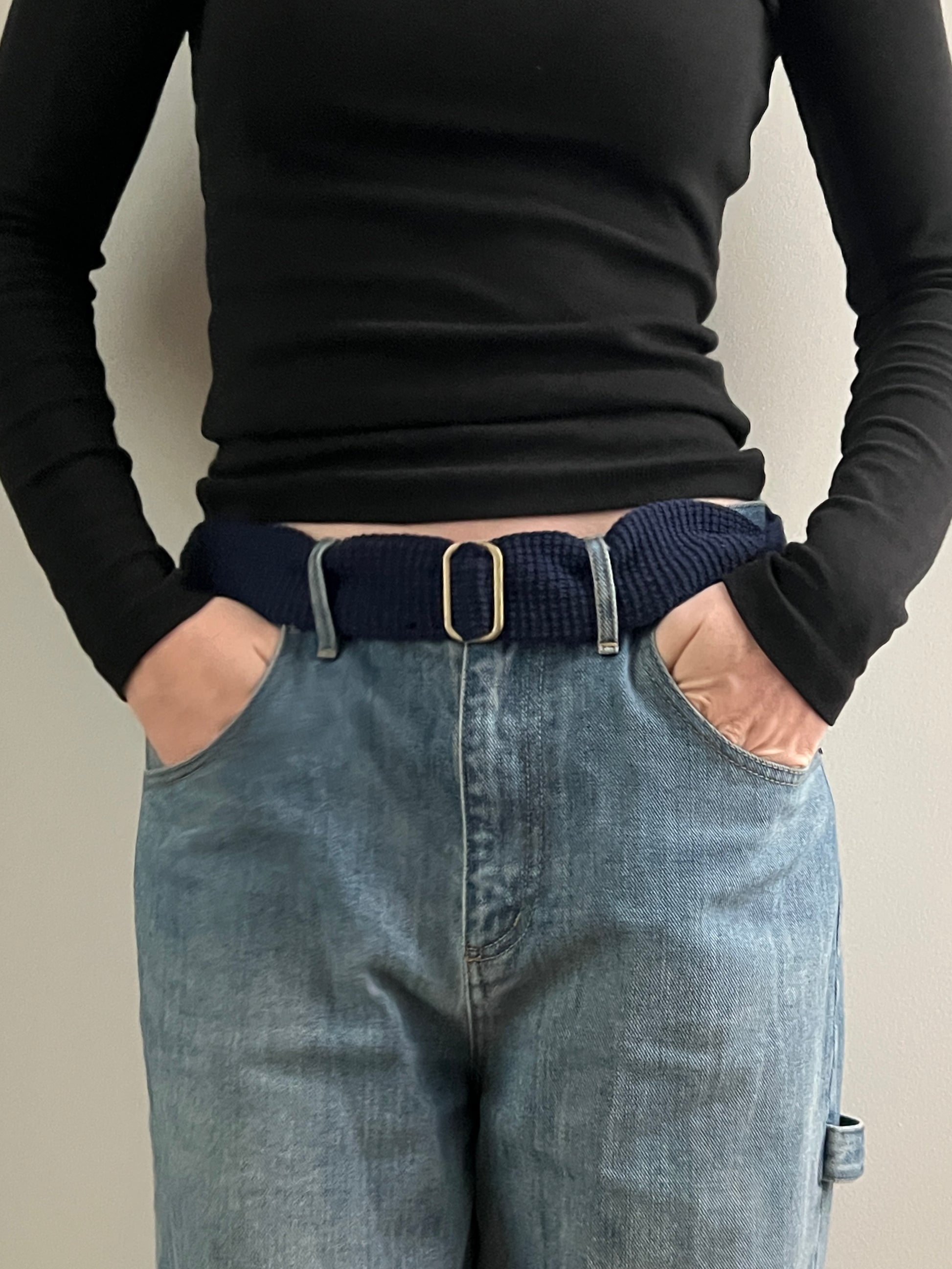 Person wearing a black long-sleeve top and blue jeans with a belt, standing against a plain background.
