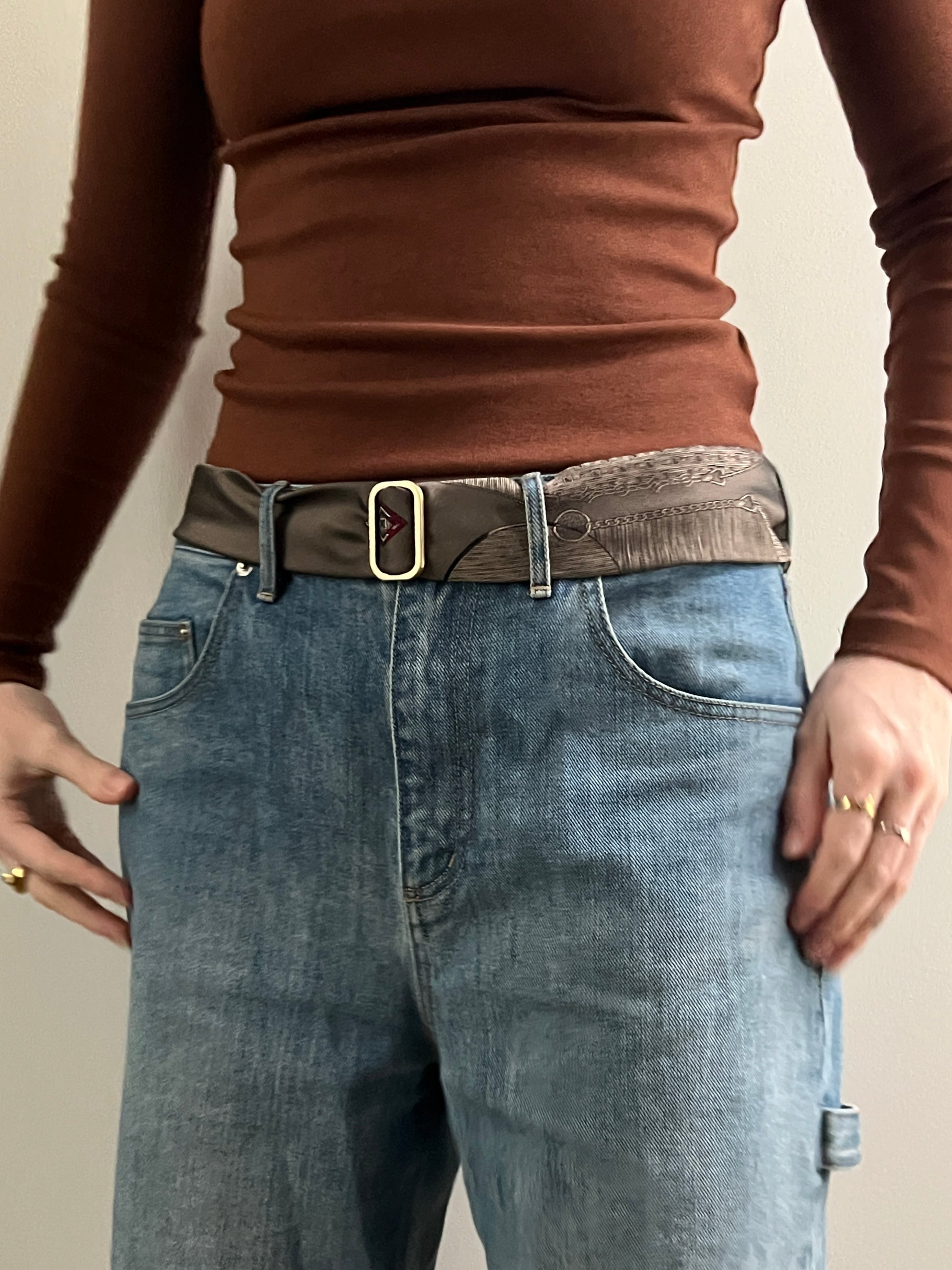 Person wearing a brown long-sleeve top and blue jeans with a belt on a neutral background