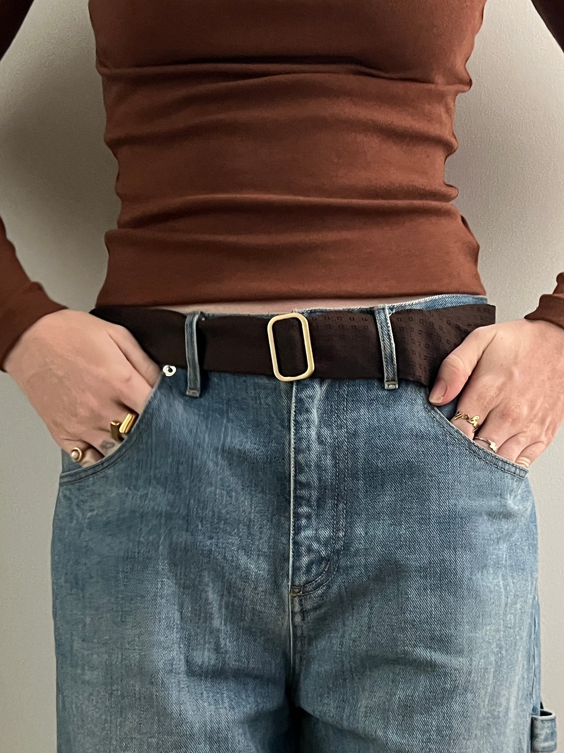 Person wearing a brown shirt and blue jeans with a belt, on a plain background