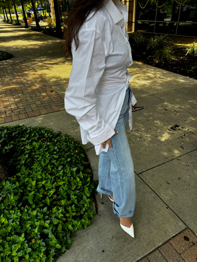 Female wearing a white cinched button down shirt and jeans standing on a sidewalk with greenery around.