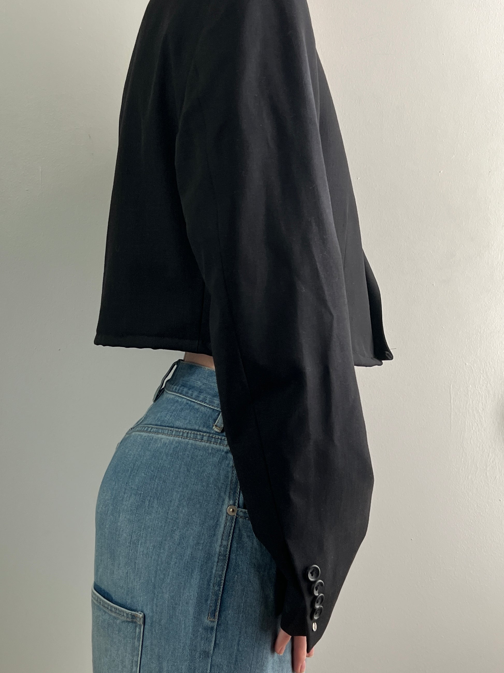 Female wearing a black cropped blazer over blue jeans on a plain background