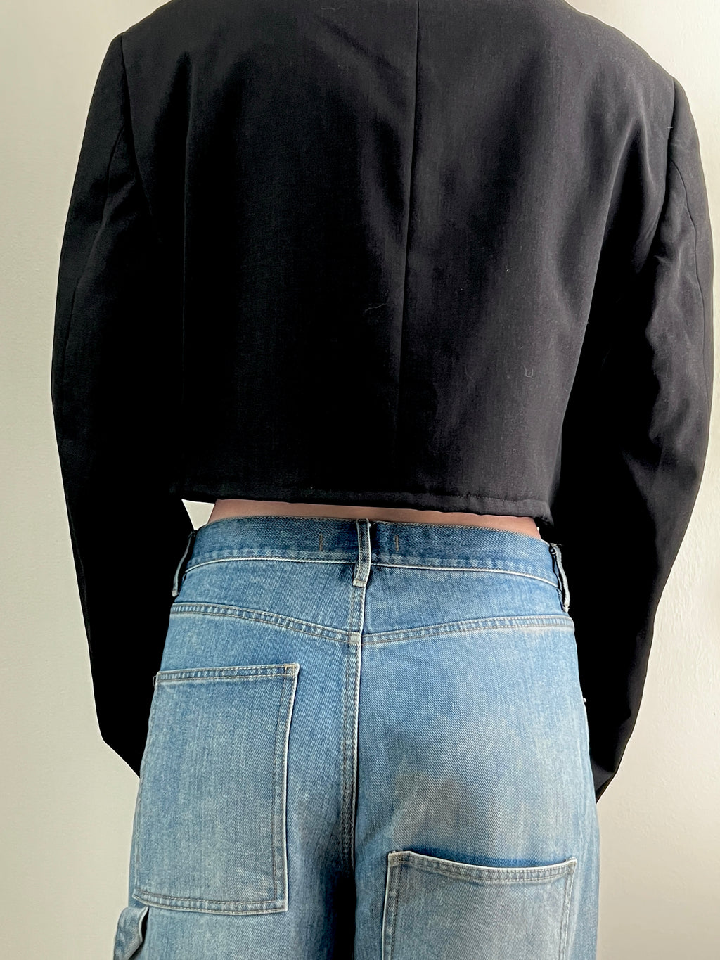 Female wearing a cropped black jacket and blue jeans from behind on a plain background
