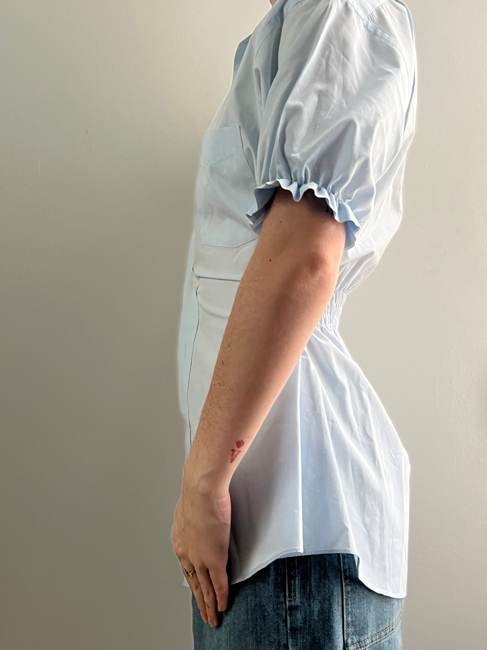 Female wearing a pale blue short sleeve puff sleeve button down blouse with a cinched back facing left.
