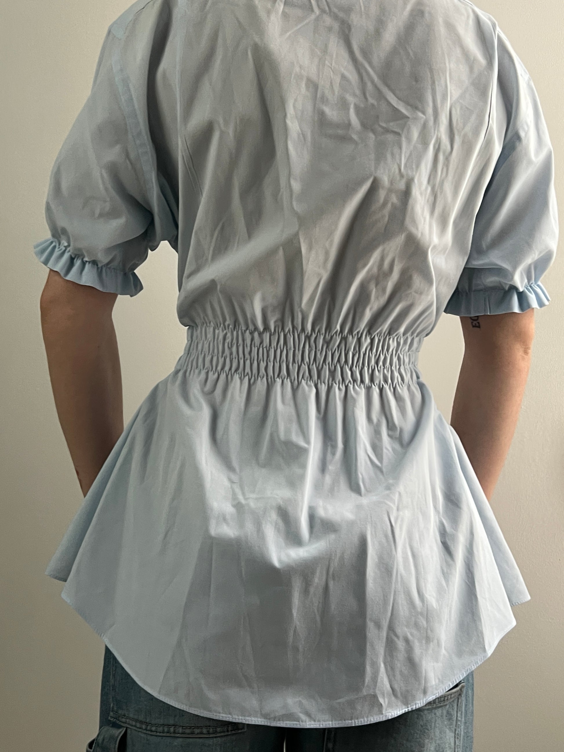 Female wearing a pale blue short sleeve puff sleeve button down blouse with a cinched back facing back.