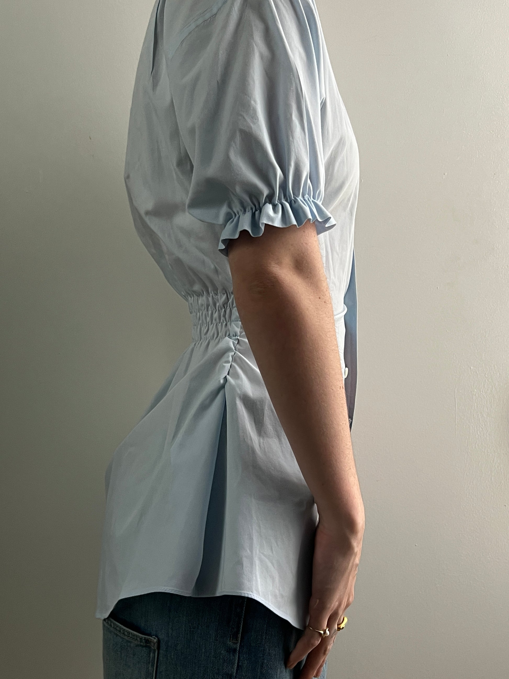 Female wearing a pale blue short sleeve puff sleeve button down blouse with a cinched back facing right.