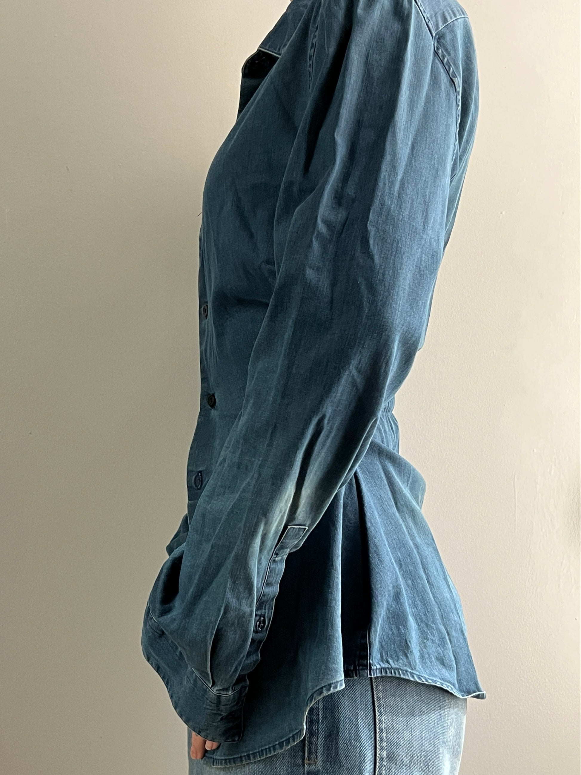 Female wearing a medium dark denim button down blouse with a cinched elastic back facing left.