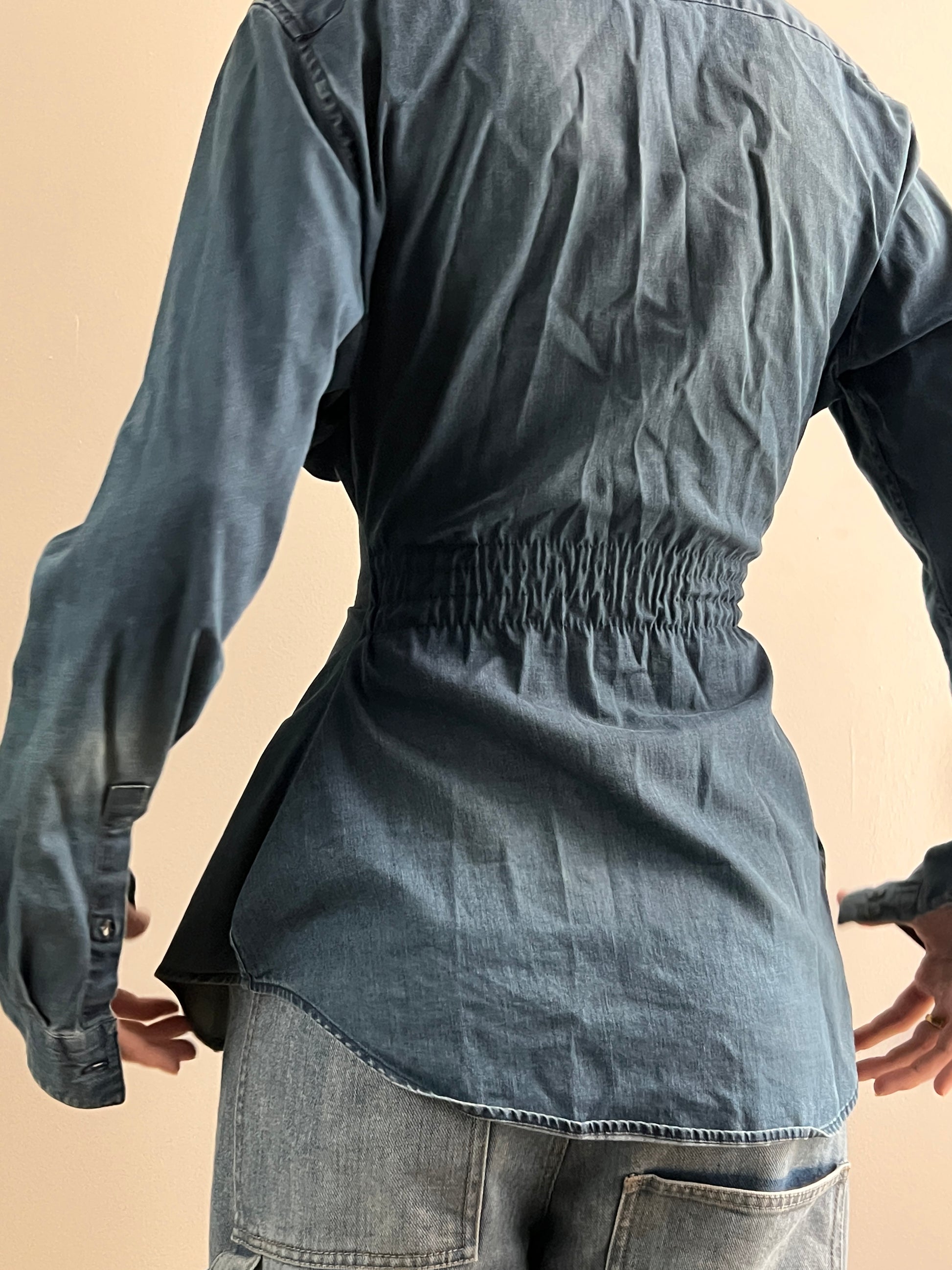 Female wearing a medium dark denim button down blouse with a cinched elastic back facing back.