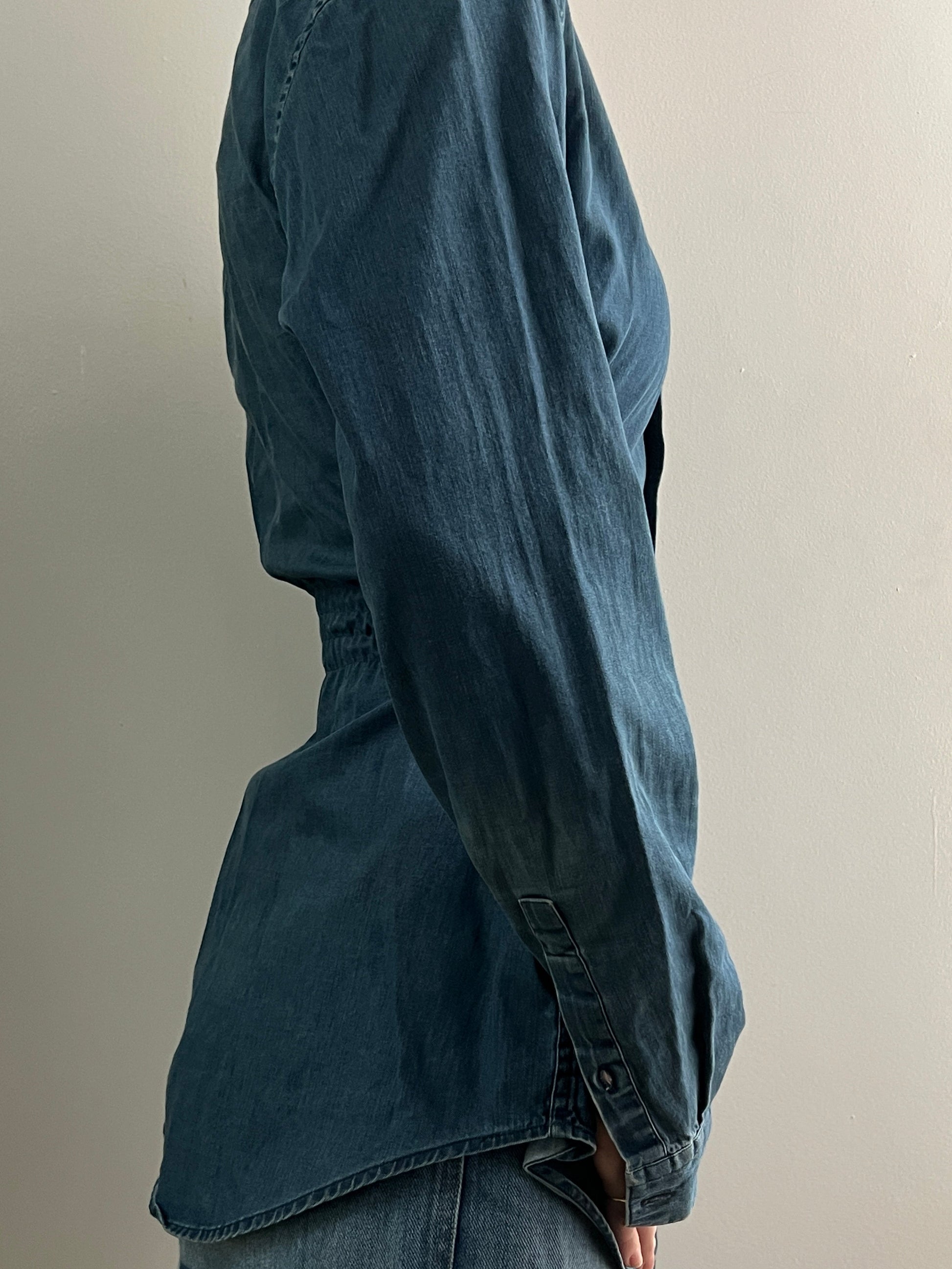 Female wearing a medium dark denim button down blouse with a cinched elastic back facing right.