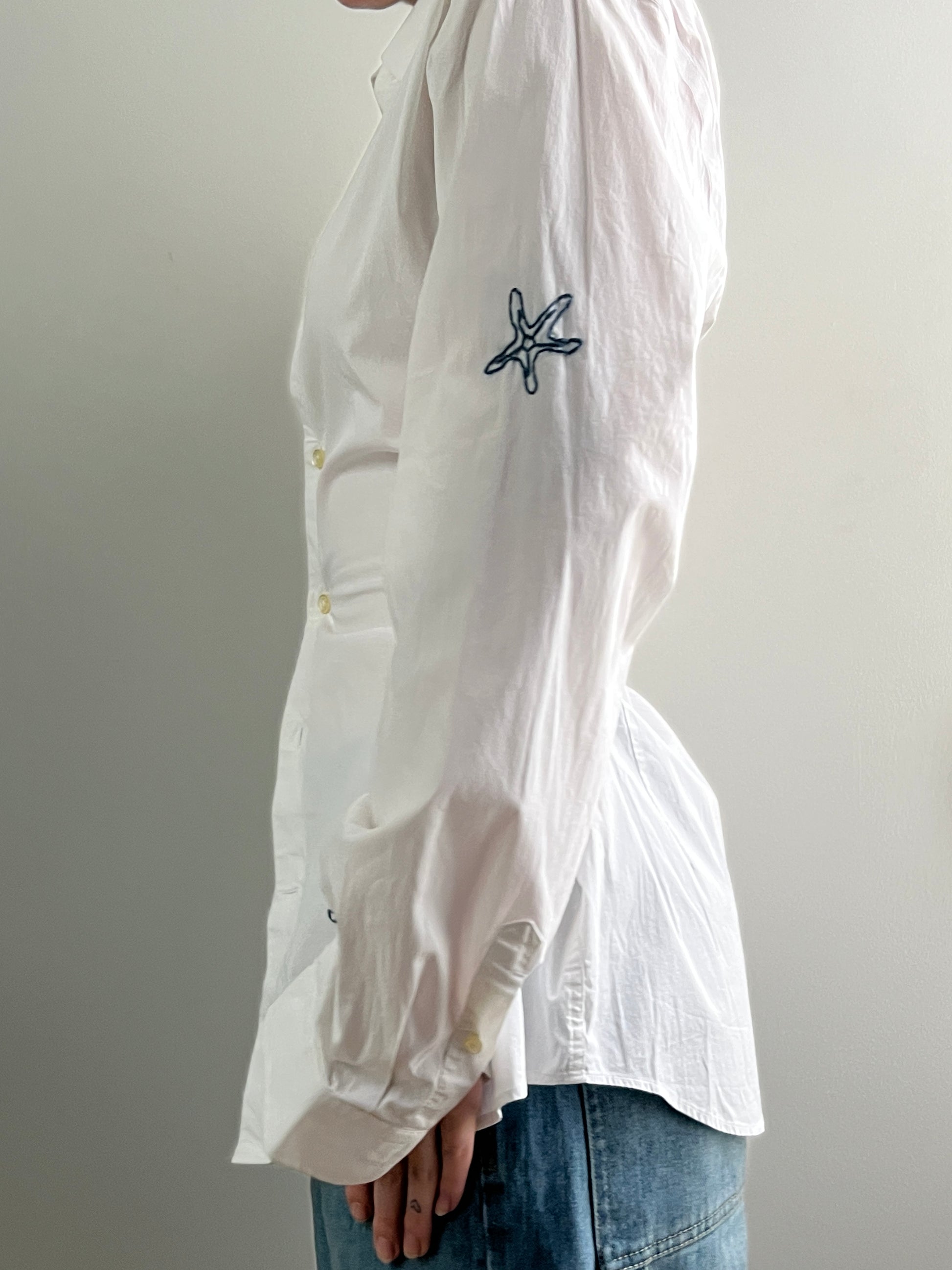 Female wearing a white navy nautically embroidered button down blouse with a cinched elastic back facing left.