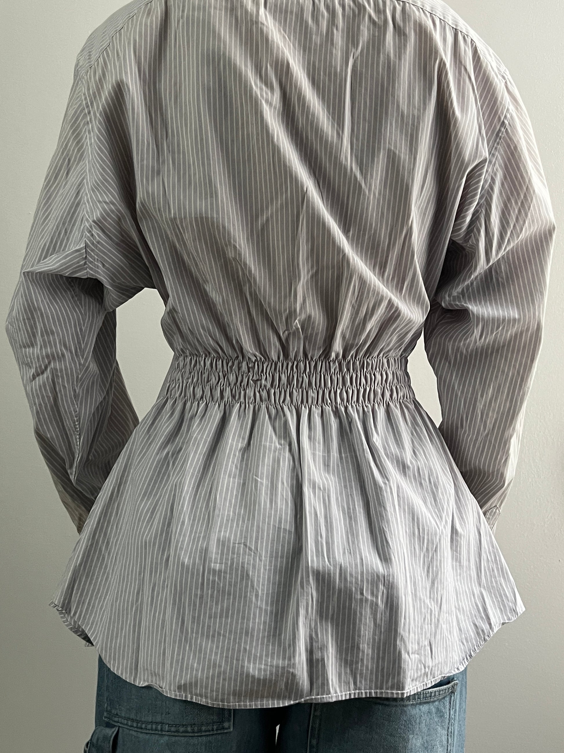 Female wearing a gray striped button down blouse with a cinched elastic back facing back.