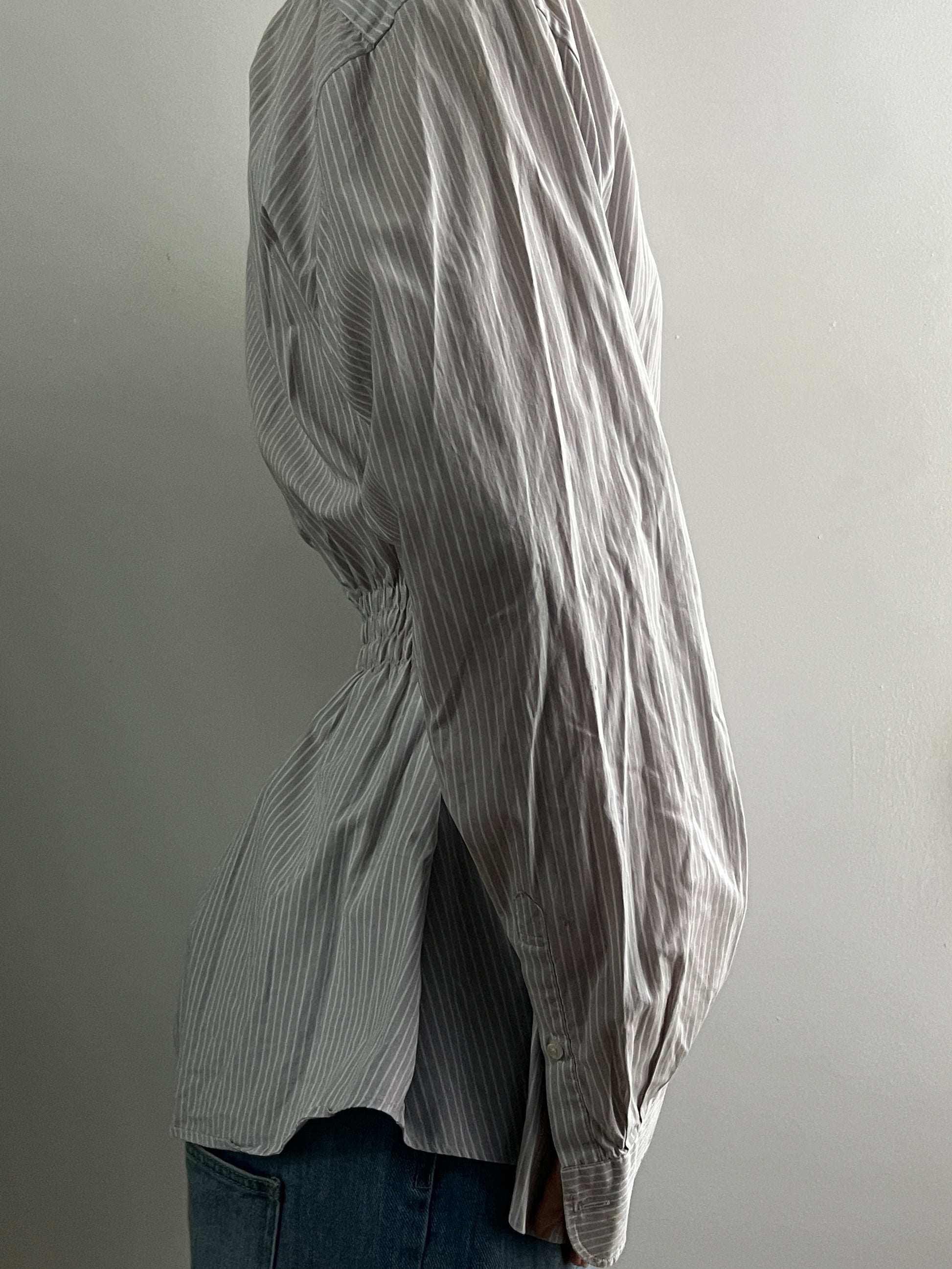 Female wearing a gray striped button down blouse with a cinched elastic back facing right.