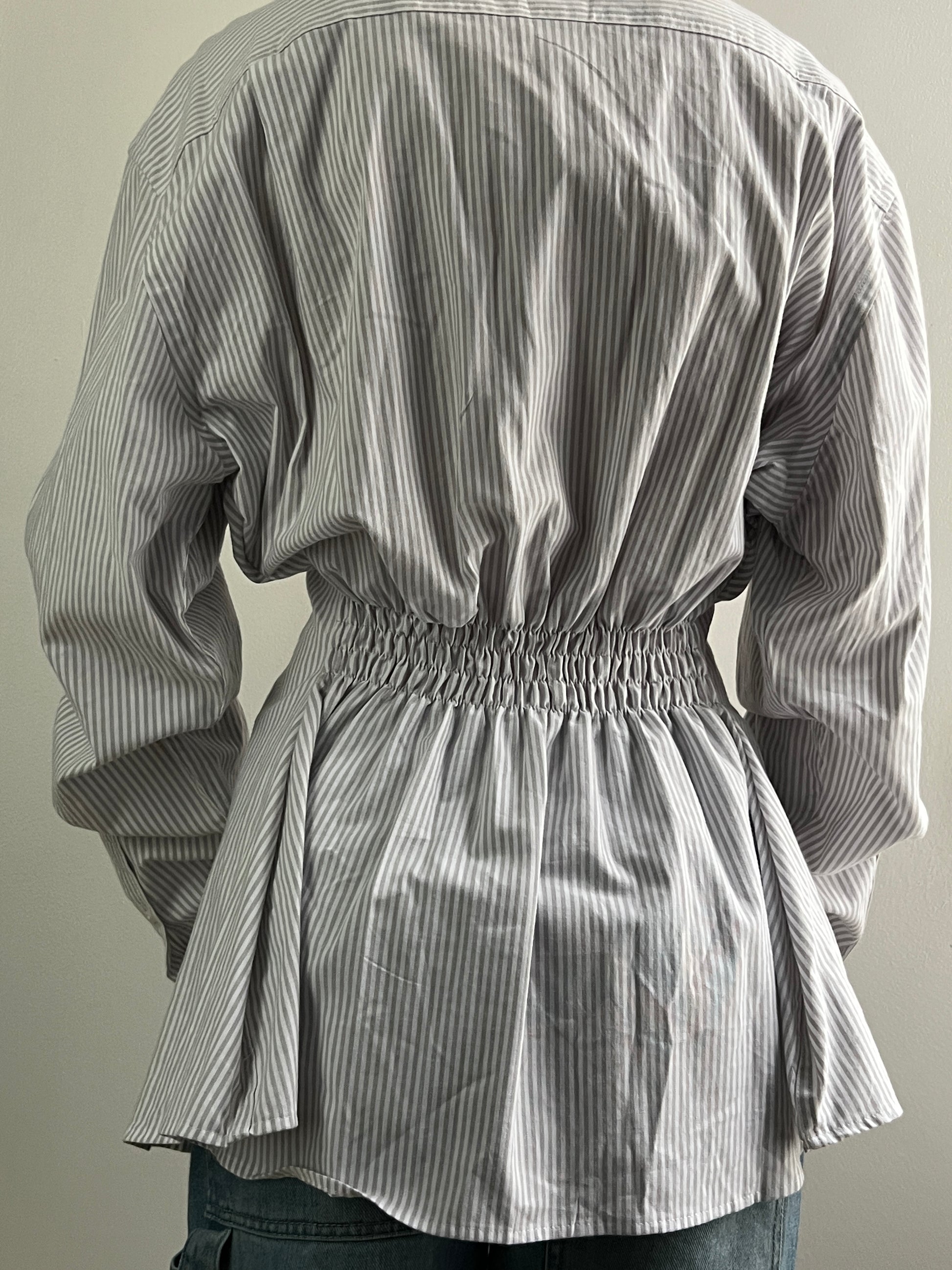 Female wearing a gray striped button down blouse with a cinched elastic back facing back.