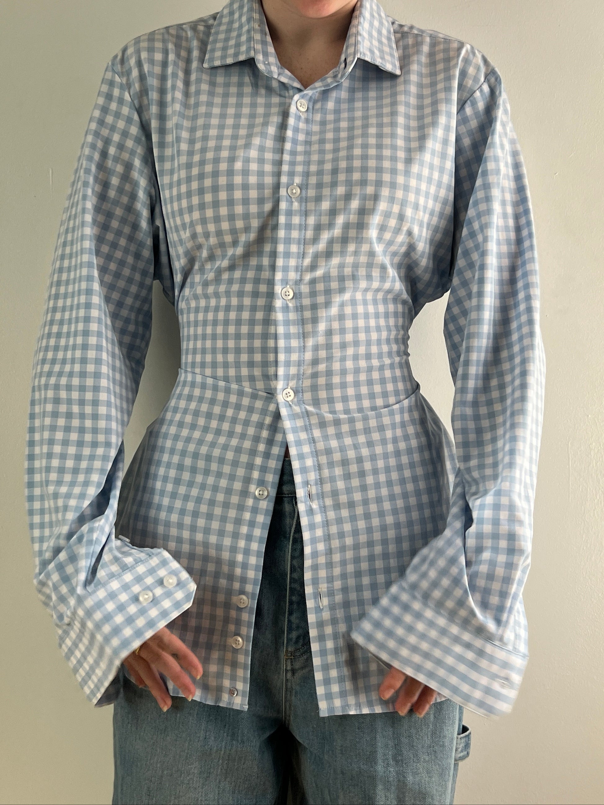 Female wearing a baby blue gingham button down blouse with a cinched elastic back facing front.