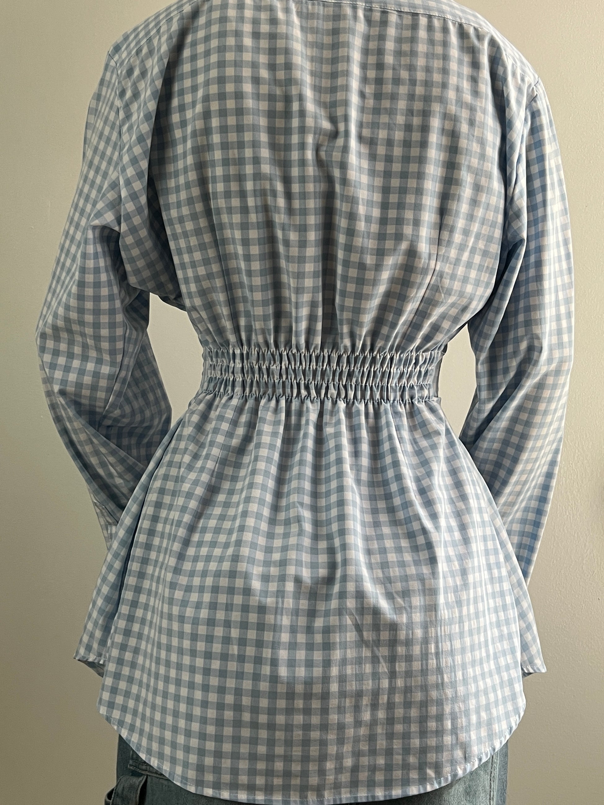 Female wearing a baby blue gingham button down blouse with a cinched elastic back facing back.
