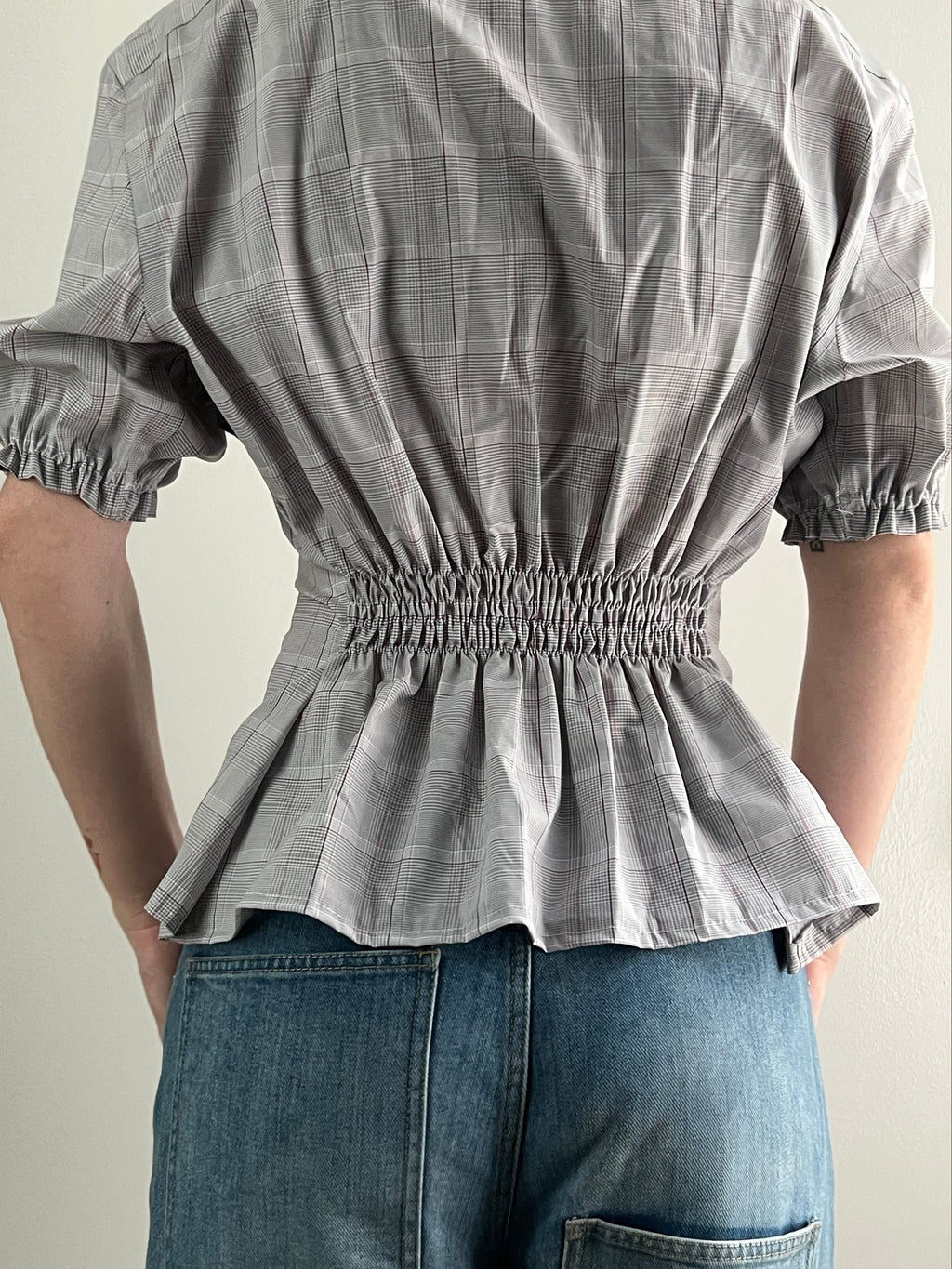 Female wearing a plaid short sleeve puff sleeve button down blouse with a cinched back facing right.