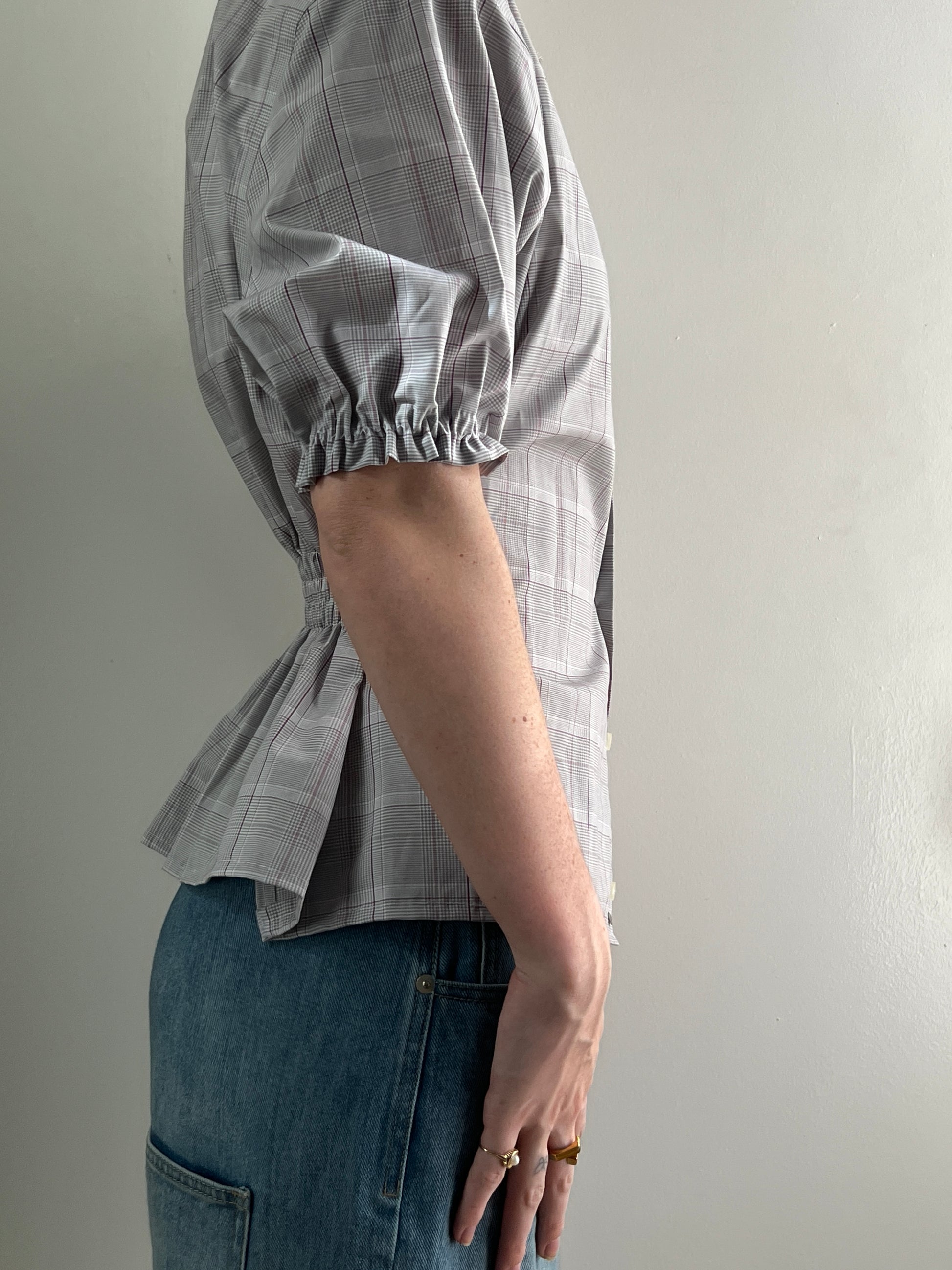 Female wearing a plaid short sleeve puff sleeve button down blouse with a cinched back facing right.
