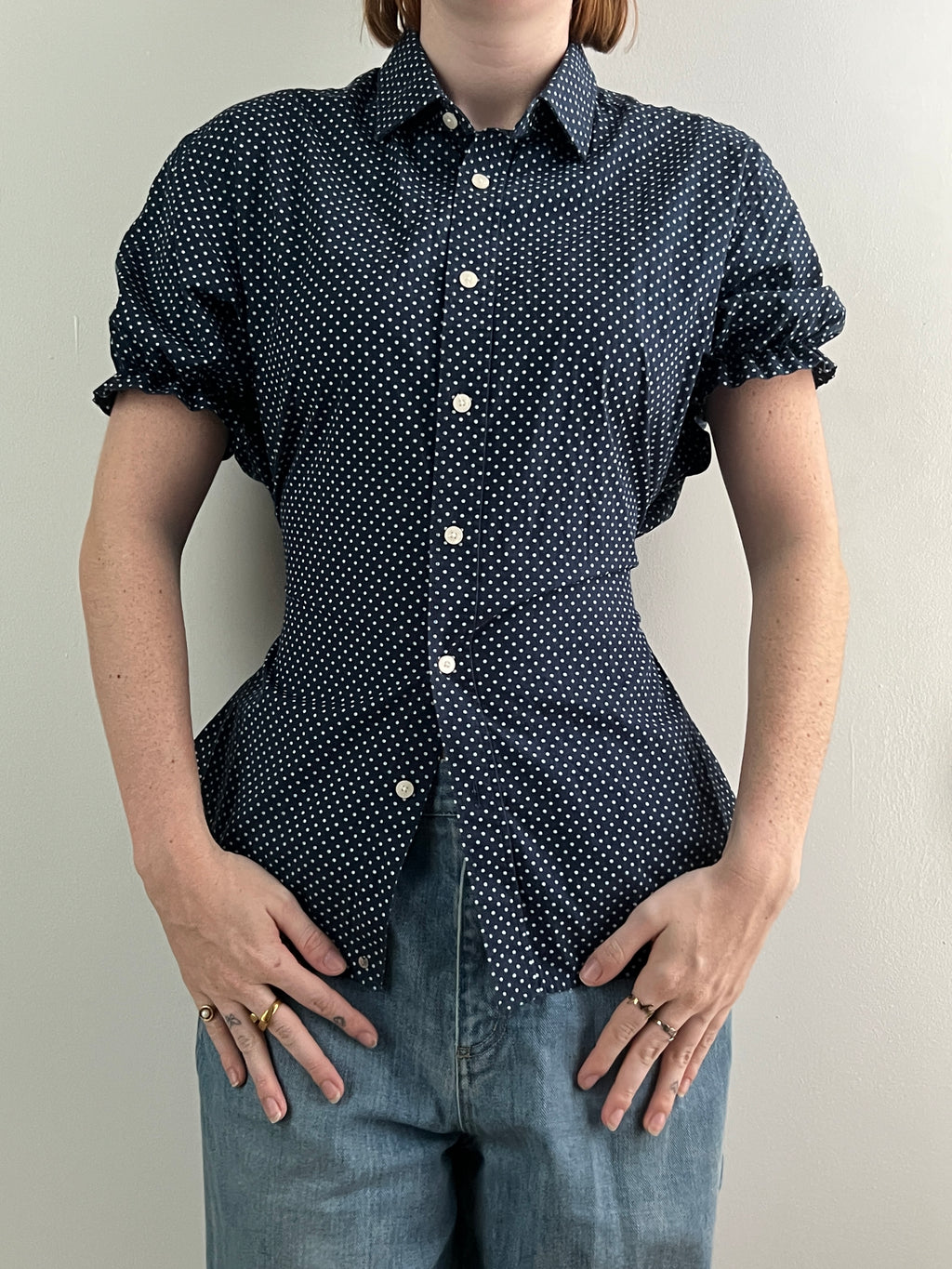 Female wearing a navy polka dot short sleeve puff sleeve button down blouse with a cinched back facing front.