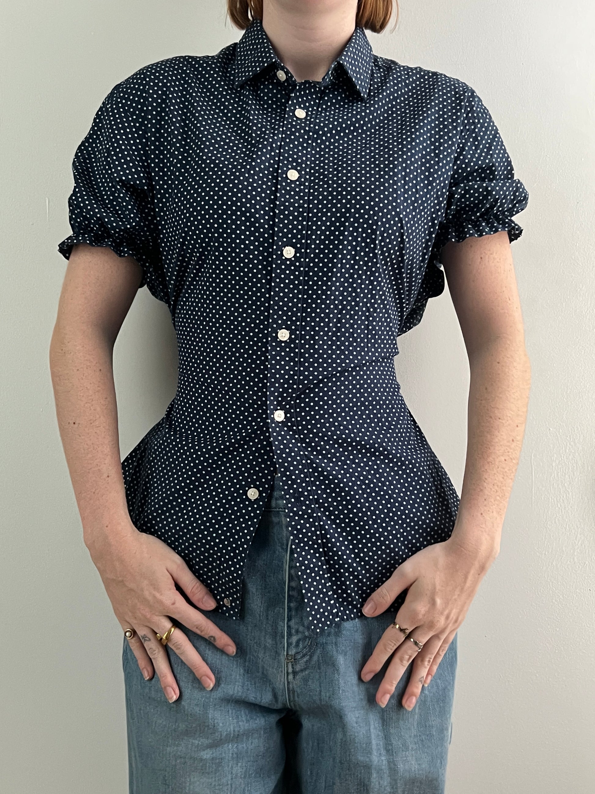 Female wearing a navy polka dot short sleeve puff sleeve button down blouse with a cinched back facing front.