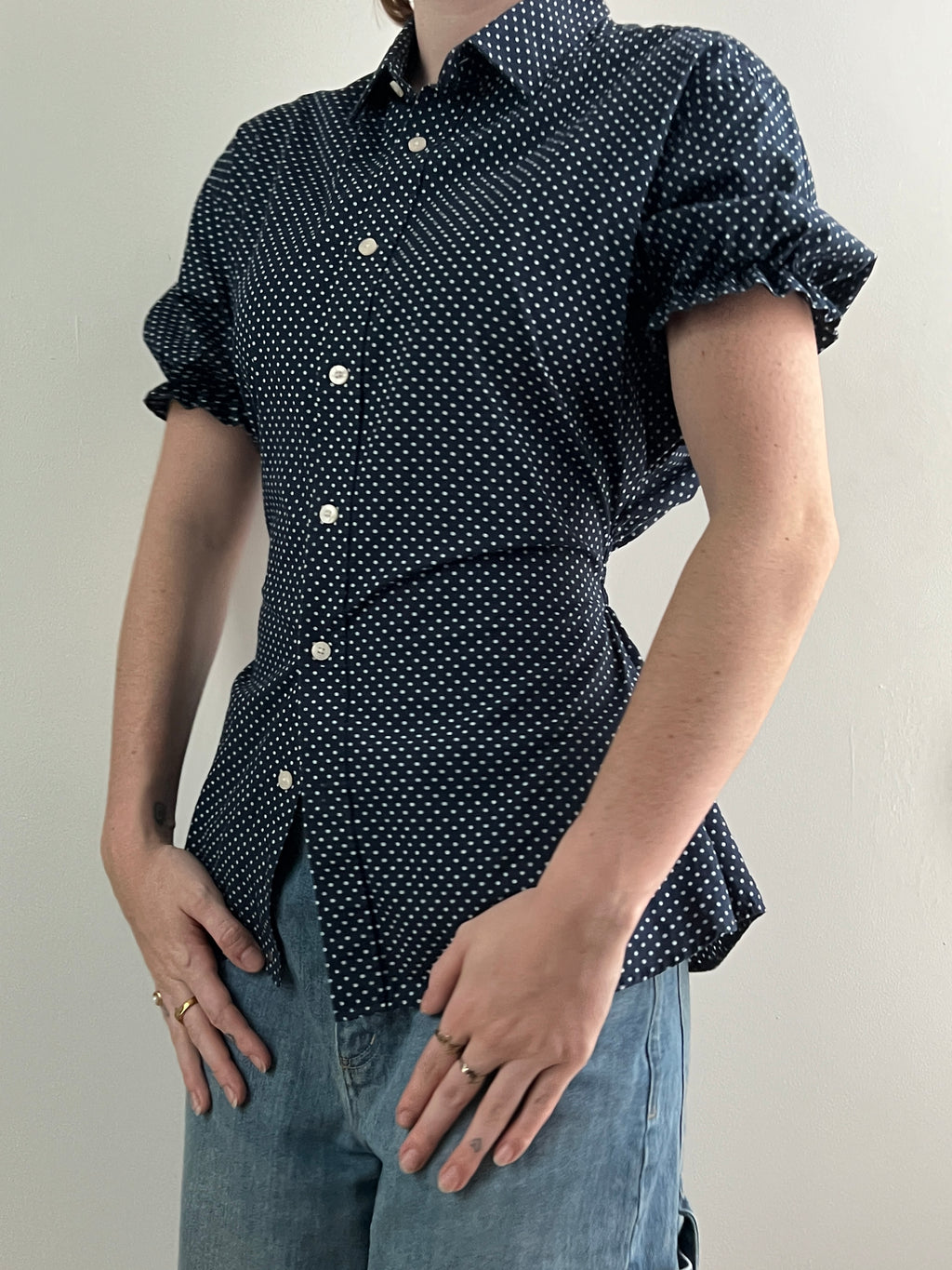 Female wearing a navy polka dot short sleeve puff sleeve button down blouse with a cinched back facing left front.
