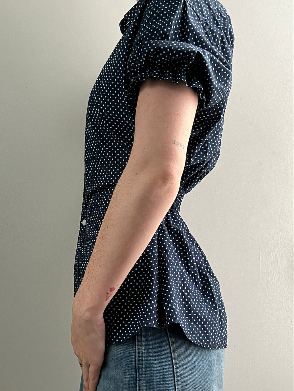 Female wearing a navy polka dot short sleeve puff sleeve button down blouse with a cinched back facing left.