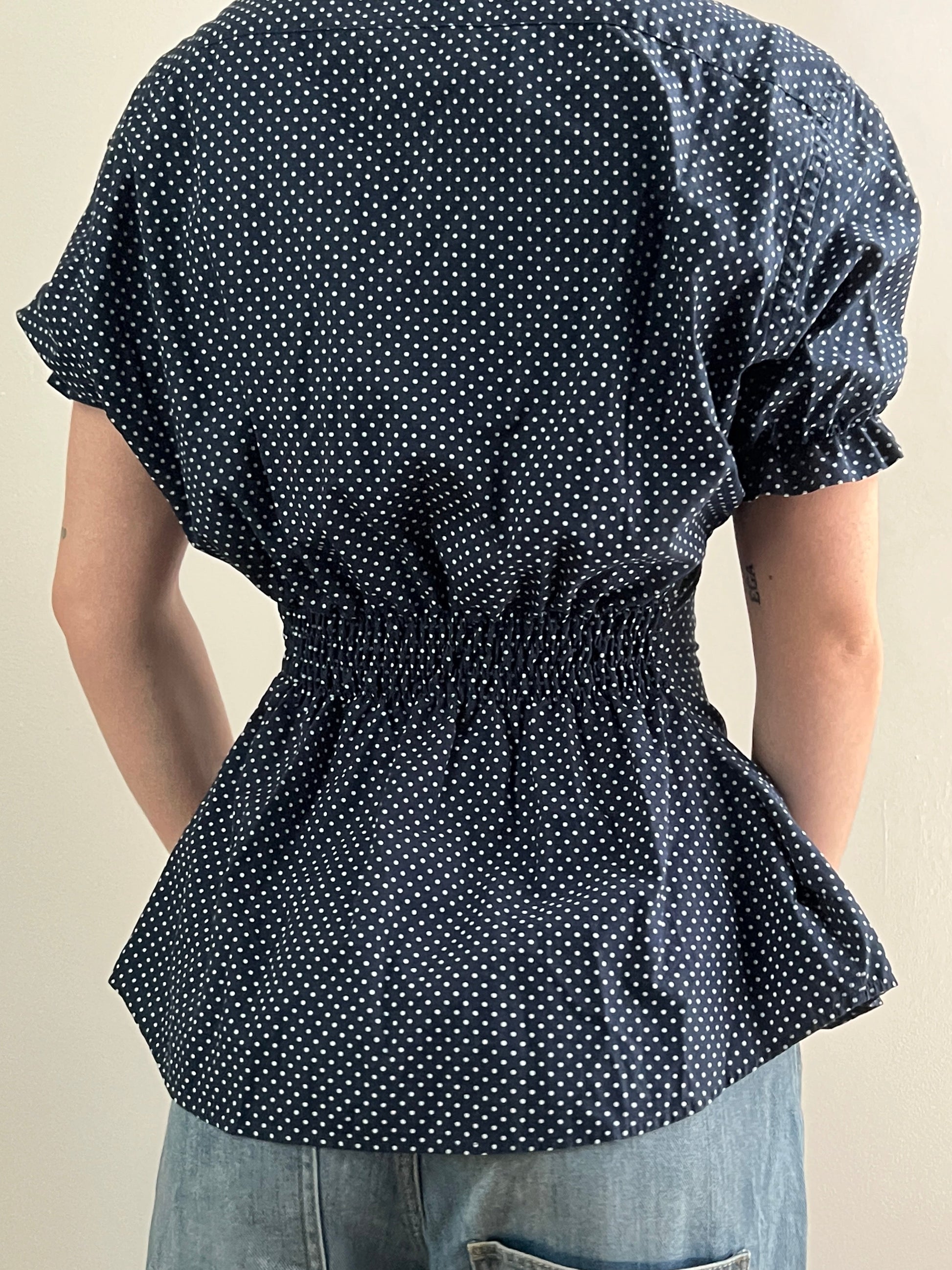 Female wearing a navy polka dot short sleeve puff sleeve button down blouse with a cinched back facing back.