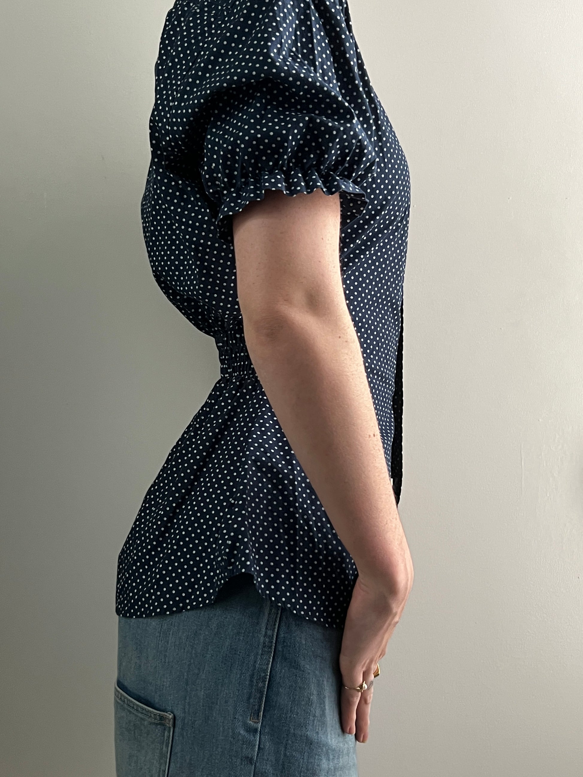 Female wearing a navy polka dot short sleeve puff sleeve button down blouse with a cinched back facing right.