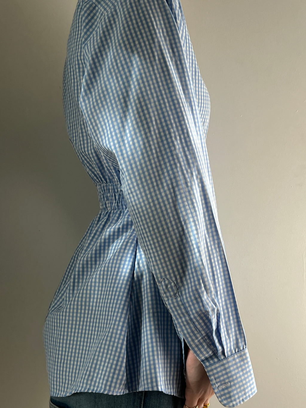 Female wearing a baby blue gingham button down blouse with a cinched elastic back facing right.
