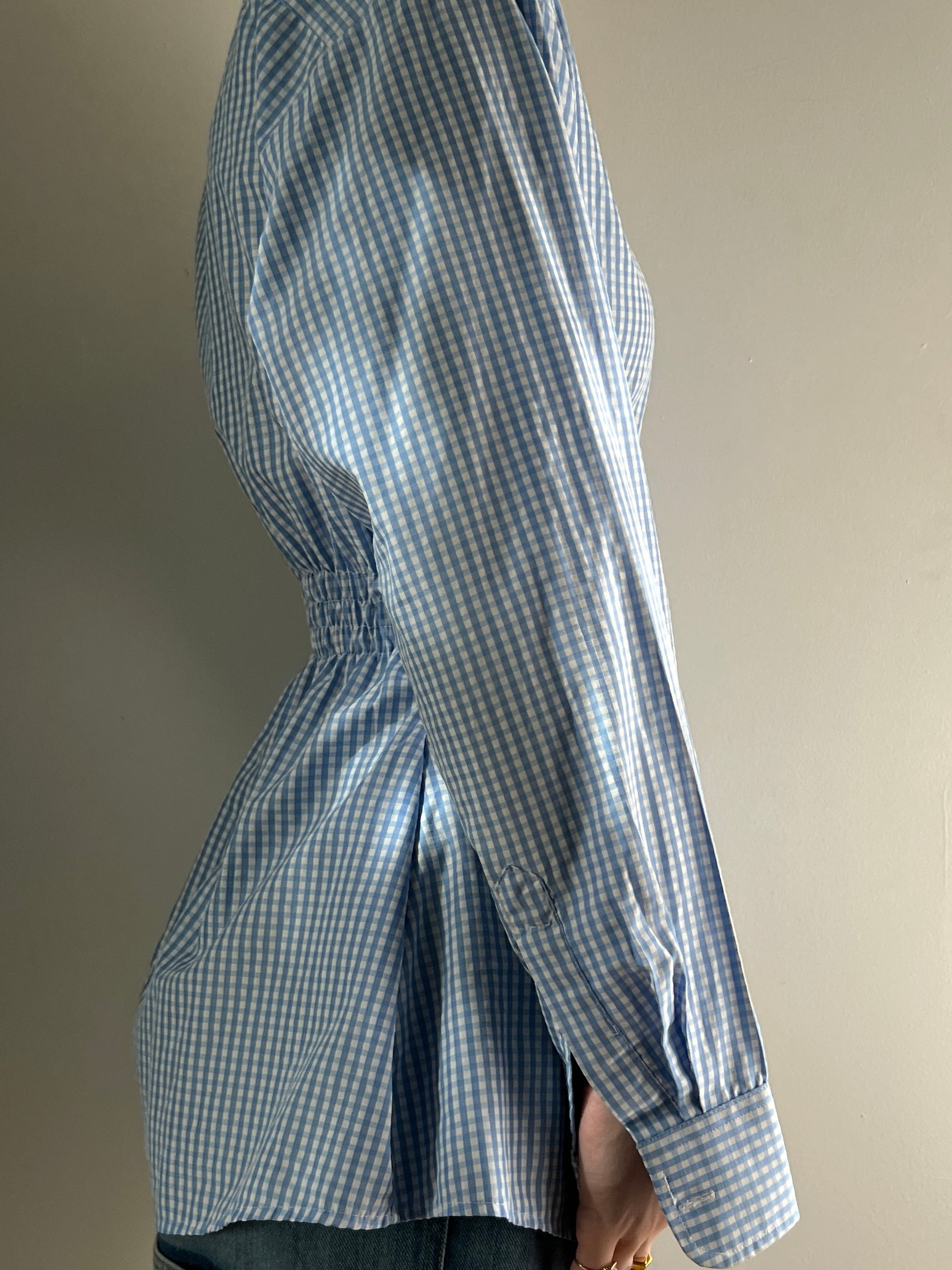 Female wearing a baby blue gingham button down blouse with a cinched elastic back facing right.